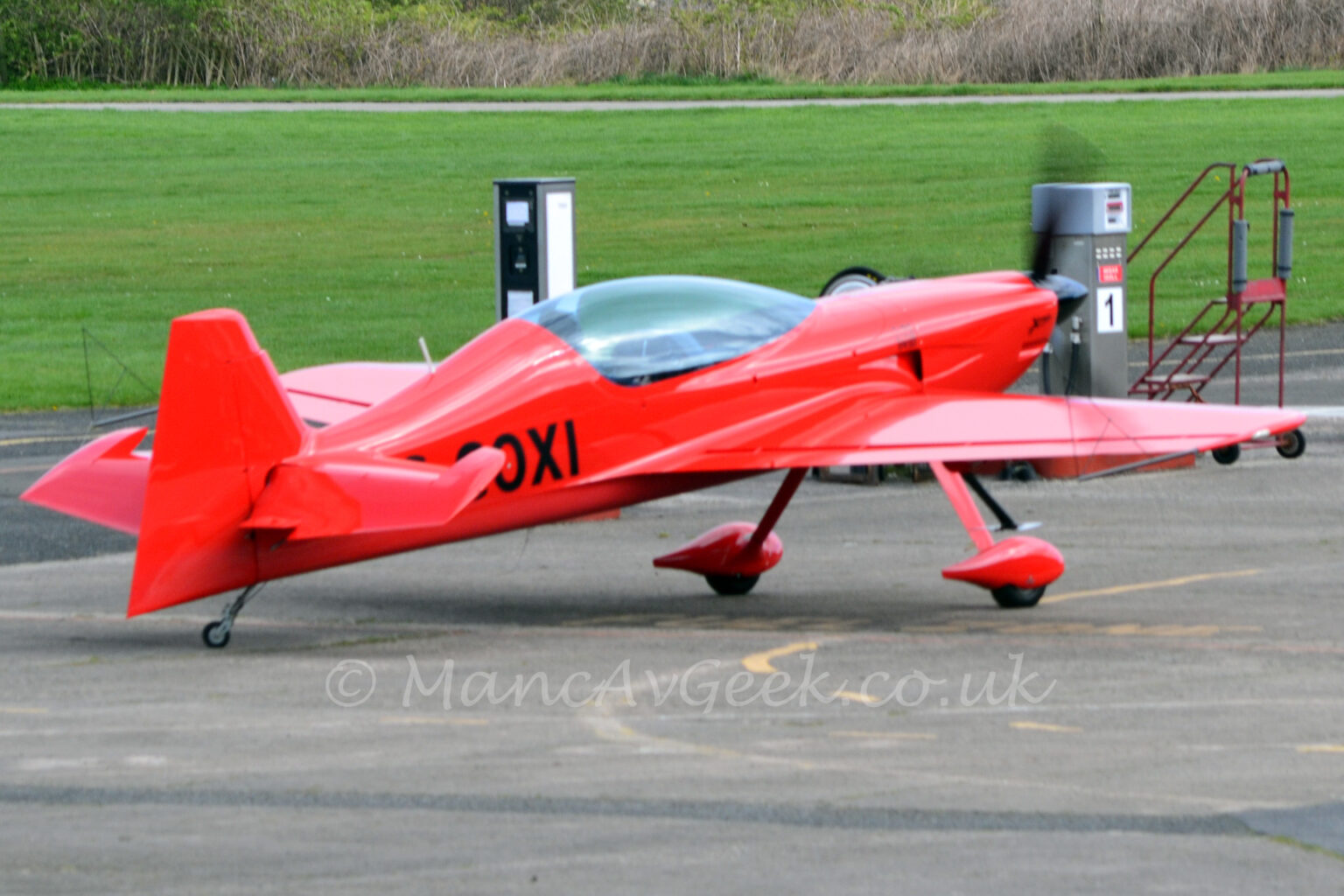 Side view of a rather sleek looking, single engined light aircraft parked taxiing from left to right along a grey tarmac taxiway. The plane is almost entirely red, with the black letters "OXI" (part of the registration) visible on the side of the rear fuselage. A large bubble canopy covers the 2 seat cockpit. Long, spindly legs coming from under the wing carry the main wheels, which have streamlined red spats over the wheels. Green grass fills most of the background, with trees stretching along the top of the frame in the distance.