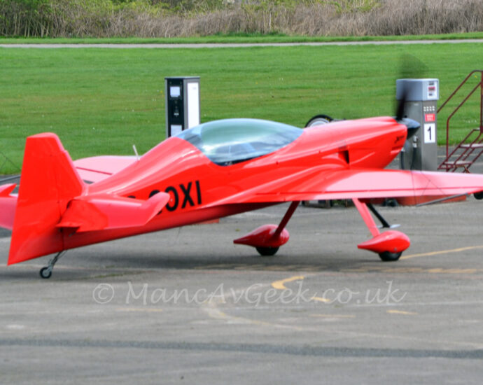 Side view of a rather sleek looking, single engined light aircraft parked taxiing from left to right along a grey tarmac taxiway. The plane is almost entirely red, with the black letters "OXI" (part of the registration) visible on the side of the rear fuselage. A large bubble canopy covers the 2 seat cockpit. Long, spindly legs coming from under the wing carry the main wheels, which have streamlined red spats over the wheels. Green grass fills most of the background, with trees stretching along the top of the frame in the distance.