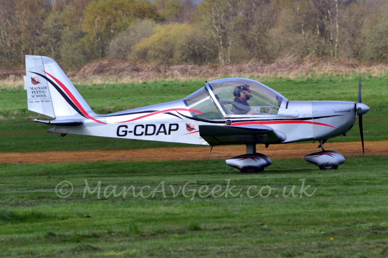 Manchester, Barton, EGCB, Evektor, EV97, MainAirFlyingSchool, AvGeek, aviation, planespotting, photography Side view of a small, single-engined light aircraft taxiing from left to right along a grass runway. The plane is in a silvery grey finish, with a pair of black and red tripes entwined around each other, arcing their way along the fuselage. Similar, smaller, stripes are on the silver spats over the nose wheel and main wheels The black registration "G-CDAP" is on the side of the lower rear fuselage. A couple of people can be seen sitting in the cockpit through the large bubble canopy. Green grass fills the foreground and most of the background, with trees and bushes marking the airfield perimeter in the distance.