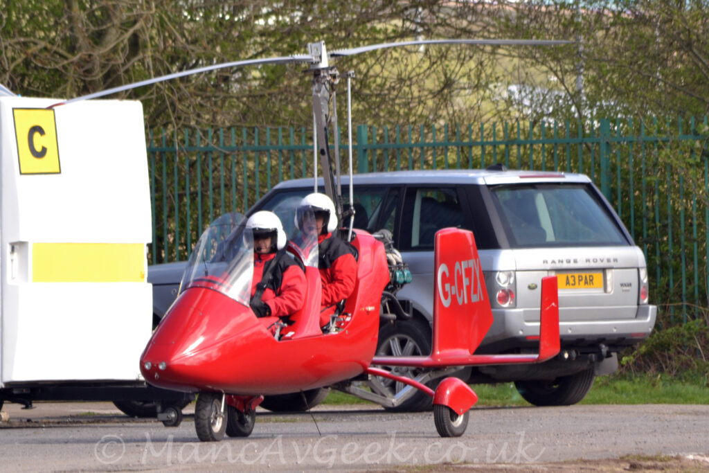 Side view of a small, red, 2-seat autogyro taxiing from right to left along a grey tarmac taxiway. The aircraft is red, with a tandem 2 seat cockpit behind a large perspex panel, with a rotor mast with a 2-bladed rotor on top coming out of the top of the fuselage behind the rear seat. A triple tail unit is on a long boom coming from the lower rear of the fuselage, with the silver registration "G-CFZX" on the sides of the larger, central tail. Both of the seats are filled, with 2 people wearing red jumpsuits and white helmets, with black straps holding them in place. A silver car with black trim is in the background immediately behind this aircraft, with a white box on wheels on the left, a yellow square with the black letter "C" in the top left corner. Trees behind a black fence fill the rest of the frame.