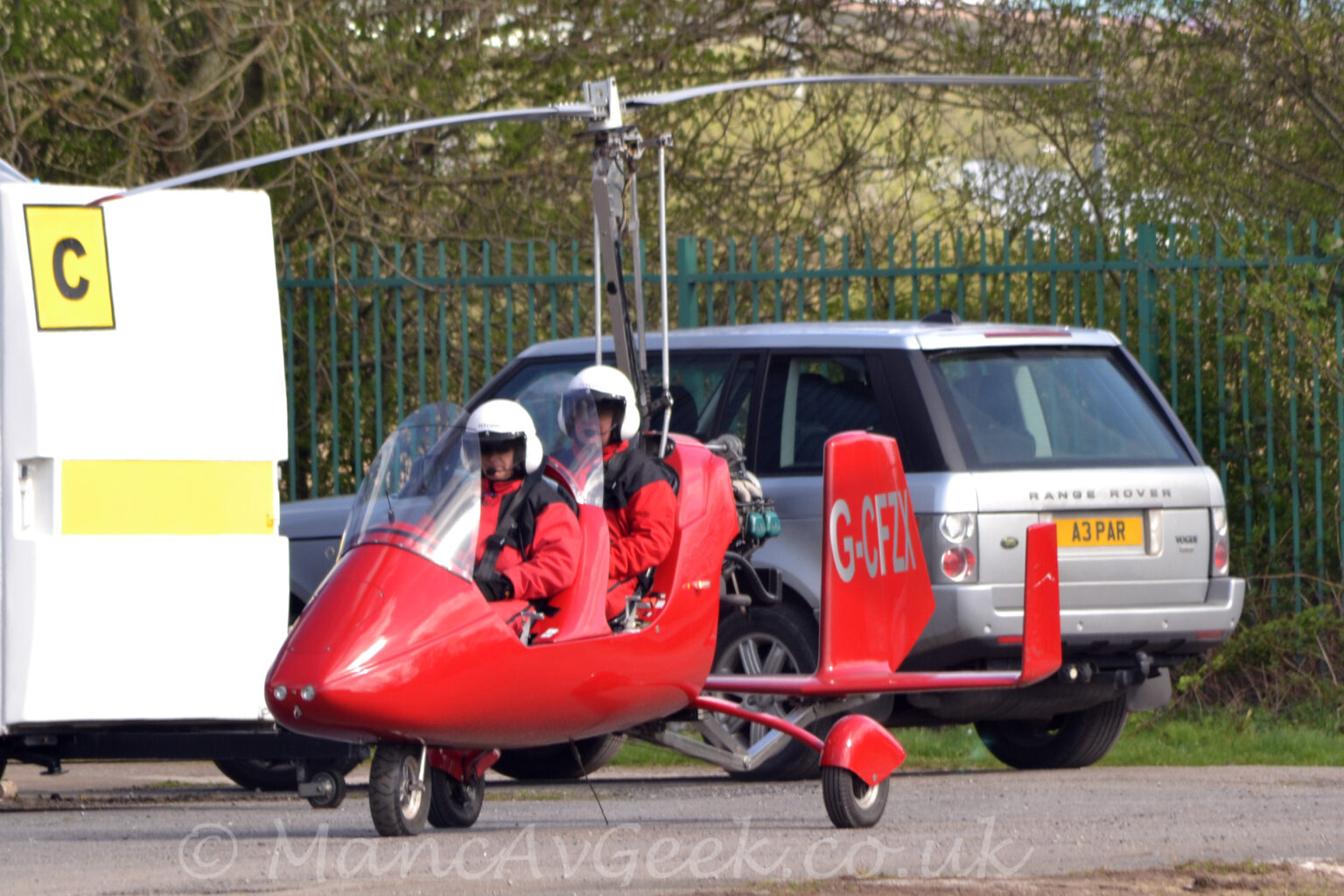 Side view of a small, red, 2-seat autogyro taxiing from right to left along a grey tarmac taxiway. The aircraft is red, with a tandem 2 seat cockpit behind a large perspex panel, with a rotor mast with a 2-bladed rotor on top coming out of the top of the fuselage behind the rear seat. A triple tail unit is on a long boom coming from the lower rear of the fuselage, with the silver registration "G-CFZX" on the sides of the larger, central tail. Both of the seats are filled, with 2 people wearing red jumpsuits and white helmets, with black straps holding them in place. A silver car with black trim is in the background immediately behind this aircraft, with a white box on wheels on the left, a yellow square with the black letter "C" in the top left corner. Trees behind a black fence fill the rest of the frame.
