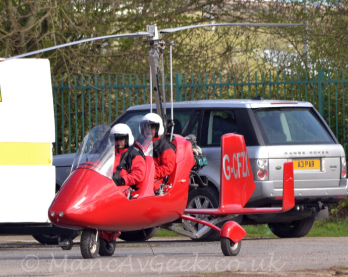 Side view of a small, red, 2-seat autogyro taxiing from right to left along a grey tarmac taxiway. The aircraft is red, with a tandem 2 seat cockpit behind a large perspex panel, with a rotor mast with a 2-bladed rotor on top coming out of the top of the fuselage behind the rear seat. A triple tail unit is on a long boom coming from the lower rear of the fuselage, with the silver registration "G-CFZX" on the sides of the larger, central tail. Both of the seats are filled, with 2 people wearing red jumpsuits and white helmets, with black straps holding them in place. A silver car with black trim is in the background immediately behind this aircraft, with a white box on wheels on the left, a yellow square with the black letter "C" in the top left corner. Trees behind a black fence fill the rest of the frame.