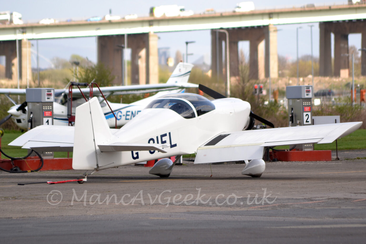 A small, single engined light aircraft parked on black tarmac, facing to the right and away from the camera. The plane is almost entirely white, with only the black registration "G-CIFL" on the side of the rear fuselage, and the large bubble canopy over the cockpit marring the whiteness. Black tarmac apron fills the foreground, with a high-winged light aircraft parked on the other side of a fuel pump in the background. A large road bridge soars into the sky in the distance, stretching across the frame, under grey sky.