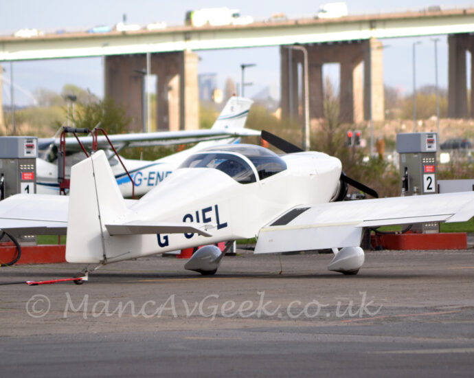 A small, single engined light aircraft parked on black tarmac, facing to the right and away from the camera. The plane is almost entirely white, with only the black registration "G-CIFL" on the side of the rear fuselage, and the large bubble canopy over the cockpit marring the whiteness. Black tarmac apron fills the foreground, with a high-winged light aircraft parked on the other side of a fuel pump in the background. A large road bridge soars into the sky in the distance, stretching across the frame, under grey sky.