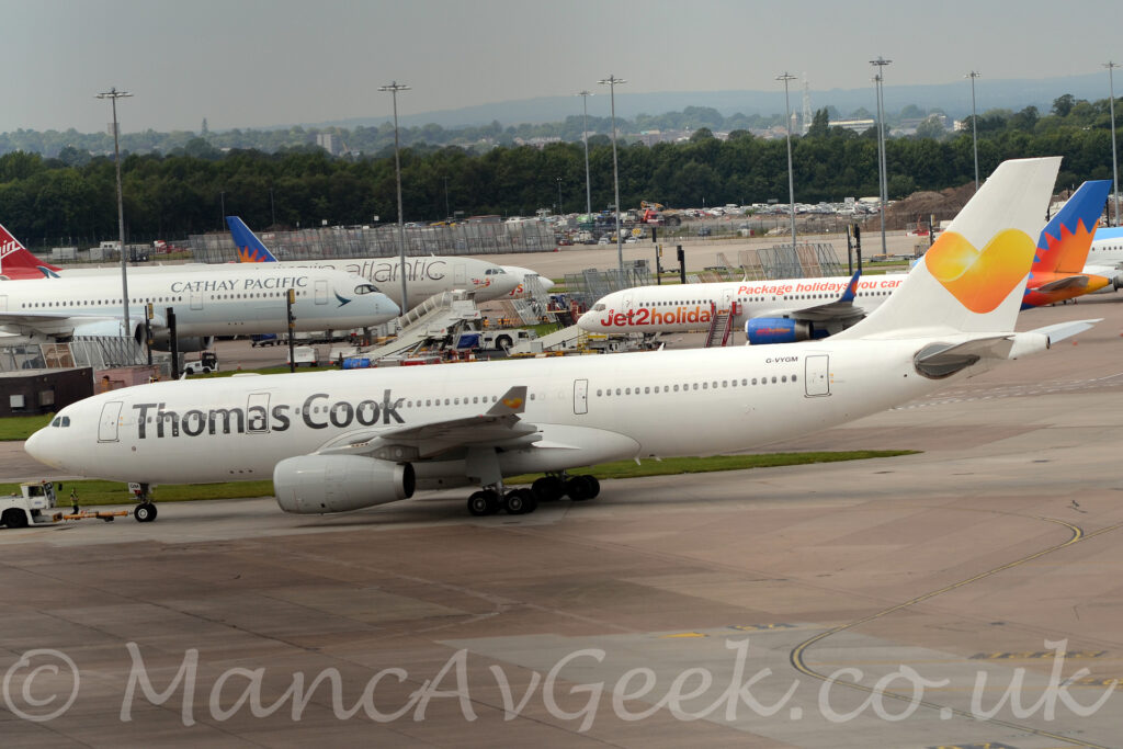 Side view of a twin engined jet airliner facing to the left but being pushed back to the right by a small white tug attached to the nose-wheel by a long yellow pole. The plane is almost entirely white, with only grey "Thomas Cook" titles on the upper forward fuselage, and the black registration "G-VYGM" on the upper rear fuselage. The white tail has a large yellow love heart in the middle, a logo repeated on the up-turned wing-tips. Grey concrete apron fills the foreground, with more concrete apron filled with more jet airliners and tall lighting poles reaching into the grey sky, with trees in the distance.