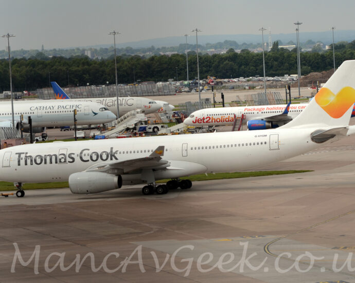 Side view of a twin engined jet airliner facing to the left but being pushed back to the right by a small white tug attached to the nose-wheel by a long yellow pole. The plane is almost entirely white, with only grey "Thomas Cook" titles on the upper forward fuselage, and the black registration "G-VYGM" on the upper rear fuselage. The white tail has a large yellow love heart in the middle, a logo repeated on the up-turned wing-tips. Grey concrete apron fills the foreground, with more concrete apron filled with more jet airliners and tall lighting poles reaching into the grey sky, with trees in the distance.