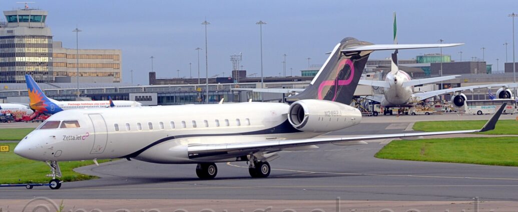 Side view of a twin engined BizJet facing to the left but being pushed back to the right by an out-of-frame tug attached to the nose wheel by a long black pole. Thew plane is mostly white, with a black stripe coming from under the front of the wing root, over the wing, then swooping up to fill the tail. There is a diagonal pink infinity symbol on the tail, a logo repeated in a smaller form under the cockpit windows, next to small "Zetta Jet" titles. Green grass at the bottom of the frame lines the side of a grey taxiway that joins another heading in to the frame in the middle. More grass lines the sides of this second taxiway, on the left and right of the frame. A terminal building stretches across the middle of the frame, with several planes parked nose in towards it, with their tails facing us, including a very large, white, 4 engined jet airliner on the right. More terminal buildings are visible above the first in the background, and a very tall building with a green-glazed tower in top on the left. Blue sky fills the rest of the frame.