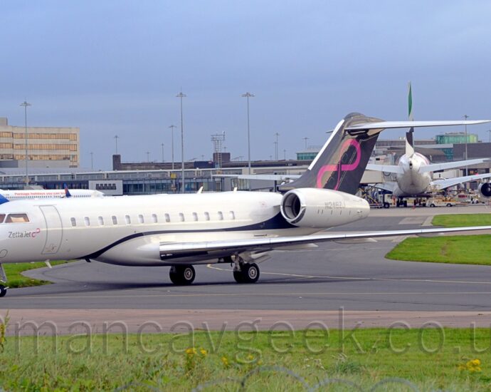 Side view of a twin engined BizJet facing to the left but being pushed back to the right by an out-of-frame tug attached to the nose wheel by a long black pole. Thew plane is mostly white, with a black stripe coming from under the front of the wing root, over the wing, then swooping up to fill the tail. There is a diagonal pink infinity symbol on the tail, a logo repeated in a smaller form under the cockpit windows, next to small "Zetta Jet" titles. Green grass at the bottom of the frame lines the side of a grey taxiway that joins another heading in to the frame in the middle. More grass lines the sides of this second taxiway, on the left and right of the frame. A terminal building stretches across the middle of the frame, with several planes parked nose in towards it, with their tails facing us, including a very large, white, 4 engined jet airliner on the right. More terminal buildings are visible above the first in the background, and a very tall building with a green-glazed tower in top on the left. Blue sky fills the rest of the frame.