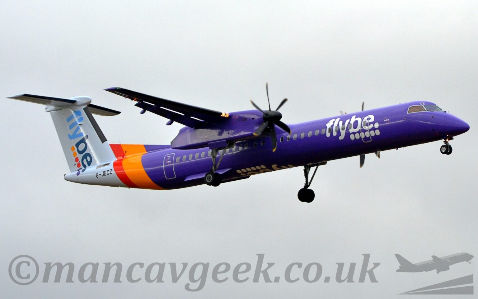 Low side view of a high-winged, twin propellor-engined airliner flying from left to right at a low altitude, with undercarriage lowered and flaps extended from the rear of the wings, suggesting it is about to land. The plane is almost entirely purple, with an orange and purple band around the rear fuselage, and a white tail-cone and tail. There are white., billboard-style "FlyBe" titles above 6 white dots on the side of the forward fuselage, with similar diagonal titles in light and dark blue on the tail, over 2 orange, 2 red, and 2 purple dots. The white titles are also inverted on the underside of the fuselage. The engine pods mounted out on the wings are also purple, with the main undercarriage hanging down on long, spindly legs from the rear. The black registration "G-JECZ" is on the side of the tail-cone right at the rear. Flat grey clouds fill the rest of the frame.