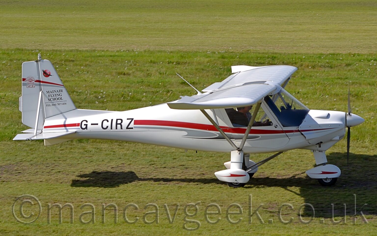 Side view of a high-winged, single-engined light aircraft taxiing from left to right along a short-mown grass taxiway. The plane is largely white, with a grey and red stripe running along the body, overlaid with the black registration "G0CIRZ" on the side of the rear fuselage. The white spats on the undercarriage has a red stripe resembling a stylised flying bird with wings outstretched for gliding. The white tail has a circular red logo for the manufacturer on the rear half, with the white text "C42" outlined in red on the bottom. The front half has the black text "MainAir Flying School", with a telephone number underneath. A red and grey stripe stretches across the whole of the tail, about 3 quarters of the way up.