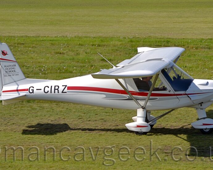 Side view of a high-winged, single-engined light aircraft taxiing from left to right along a short-mown grass taxiway. The plane is largely white, with a grey and red stripe running along the body, overlaid with the black registration "G0CIRZ" on the side of the rear fuselage. The white spats on the undercarriage has a red stripe resembling a stylised flying bird with wings outstretched for gliding. The white tail has a circular red logo for the manufacturer on the rear half, with the white text "C42" outlined in red on the bottom. The front half has the black text "MainAir Flying School", with a telephone number underneath. A red and grey stripe stretches across the whole of the tail, about 3 quarters of the way up.