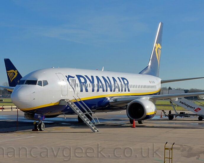 Side view of a twin engined jet airliner parked on grey concrete apron, facing to the left. The plane is largely white, with a dark blue belly, and a yellow stripe running along the body, separating the 2 colours. There are large, blue, billboard-style "RyanAir" titles on the sides of the forward fuselage, next to a blue, stylised winged Irish harp. The black registration "EI-EPA" is on the upper rear fuselage, next to a small Irish flag. The engine pods hanging under the wings are the same colours as the body - white on top, and dark blue at the bottom, separated by a yellow stripe. Built-in airstairs are deployed below the closed forwards cabin door, while white mobile airstairs are parked by the closed rear doors. Grey concrete apron fills the foreground, with a wide strip of grass leads up to a runway in the background, with trees beyond that. Pale blue sky with wisps of thin grey cloud fill the top half of the image.