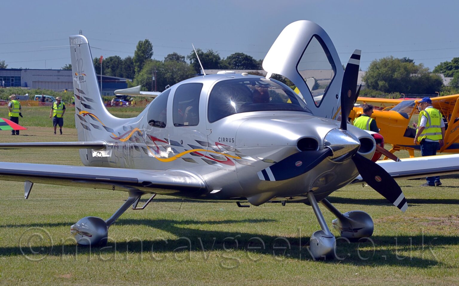 Front-side view of a single engined light aircraft parked on grass facing to the right. The plane is in a metallic grey livery, with a series of short, silver stripes running along the body, with yellow and red ribbons intertwined with and spinning through them. The silver registration "ZS-SUB" is on the lower rear fuselage. The cockpit door on the other side is open, hinged forwards and upwards. The foreground is filled with short green brass, with more grass in the background leading up to trees in the distance. Several planes are visible in the background, mostly a bright yellow plane on the right, with various wings and tails coming in to view. Several people wearing yellow hi-viz vests are scattered around the airfield, acting as marshallers for all the visitors.