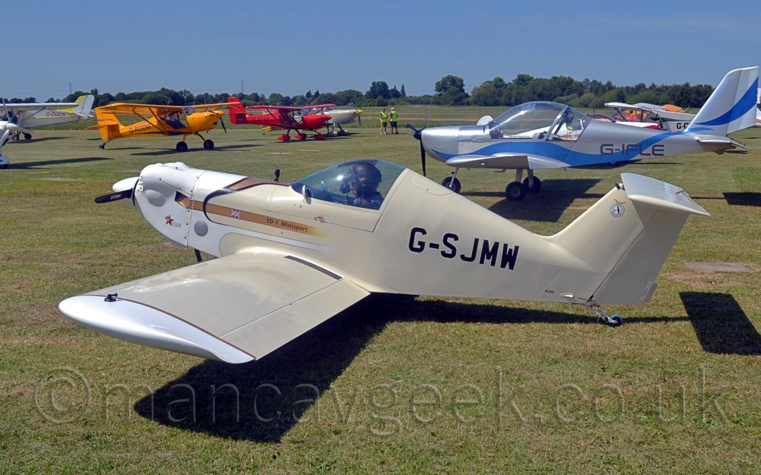 Side view of a small, single-engined, single-seat light aircraft, parked facing to the left on grass. The plane is mostly cream coloured, with white engine cowling and #wingtips, and the black registration "G-SJMW" on the rear fuselage. There is a gold stripe on the forward fuselage, with a small UK flag, and the black text "SD-1 Minisport". Strong sunlight coming from the top left of the frame is casting a strong shadow on the ground under the plane. Green grass extends into the distance, with several light aircraft parked on both sides of the image, facing in towards the middle. Trees in the distance meet blue sky with a slight hint of haze.