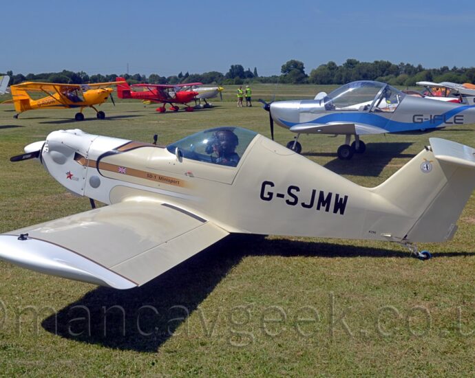 Side view of a small, single-engined, single-seat light aircraft, parked facing to the left on grass. The plane is mostly cream coloured, with white engine cowling and #wingtips, and the black registration "G-SJMW" on the rear fuselage. There is a gold stripe on the forward fuselage, with a small UK flag, and the black text "SD-1 Minisport". Strong sunlight coming from the top left of the frame is casting a strong shadow on the ground under the plane. Green grass extends into the distance, with several light aircraft parked on both sides of the image, facing in towards the middle. Trees in the distance meet blue sky with a slight hint of haze.