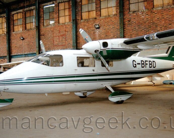 Side view of a white, high-winged, twin propellor-engined light aircraft, parked facing to the left in a brown brick hangar. The plane has a pair of green stripes running along the body, widening as they move rearwards, and a green tail outlined in white. The fixed undercarriage has spats covering the top of the wheels, with the top half painted green, the lower half in white. The registration "G-BFBD" is on the rear fuselage in black. Several light aircraft can be seen parked in the background on the left, behind this plane's nose. Brown brick walls stretch across the background, with grey vertical metal support beams at intervals. Boarded-up windows fill the top of the walls.