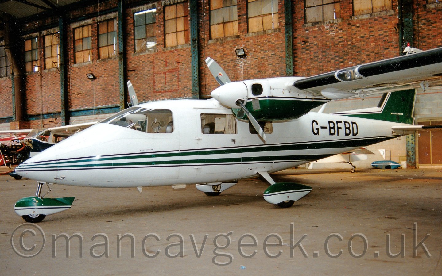 Side view of a white, high-winged, twin propellor-engined light aircraft, parked facing to the left in a brown brick hangar. The plane has a pair of green stripes running along the body, widening as they move rearwards, and a green tail outlined in white. The fixed undercarriage has spats covering the top of the wheels, with the top half painted green, the lower half in white. The registration "G-BFBD" is on the rear fuselage in black. Several light aircraft can be seen parked in the background on the left, behind this plane's nose. Brown brick walls stretch across the background, with grey vertical metal support beams at intervals. Boarded-up windows fill the top of the walls.