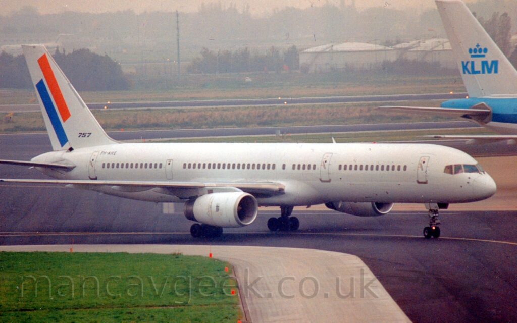 Side view of a twin engined jet airliner taxiing from left to right, following a yellow line marked on a black taxiway. The plane is almost entirely white, with only the black registration "PH-AHE" on the upper rear fuselage, with the last 2 letters repeated on the open nose wheel doors. There are a pair of diagonal stripes, one orange, the other blue, on the tail, with the black number "757" at the base. Green grass in the bottom left corner lines a black taxiway, withy more taxiways, grass, and a runway in the background leading up to domed white fuel tanks and trees in the distance under dreary grey sky.