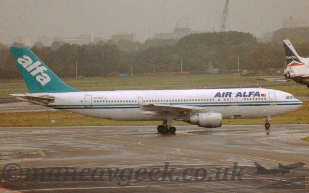 Side view of a twin engined jet airliner taxiing from left to right. The plane is mostly white, with a muted green and dark blue stripe running along the body, and dark blue "Air Alfa" titles next to a small Turkish flag on the upper forward fuselage. The black registration "TC-ALS" is on the upper rear fuselage. The tail is the same muted green, with large, white, diagonal "alfa" titles. A large expanse of grey concrete apron fills the foreground, with an equally large grassed area leads to trees in the distance, under drab grey sky. The tail of a white and black, 3 engined jet airliner can be seen on the left of the frame.