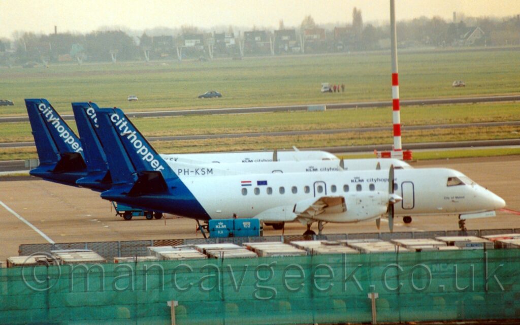 Side view of a trio of small, twin propellor-engined airliners parked next to each other, all facing to the right. The nearest plane is mostly white, with dark blue "KLM Cityhopper" titles on the upper forward fuselage, and small Dutch and EU flags above the rear cabin windows on the upper rear fuselage. The black registration "PH-KSM" is on a white fillet in front of the tail. The tail itself is dark blue, with diagonal white "Cityhopper" titles along the leading edge. The 2 planes behind it are in pretty much identical liveries. A range of luggage containers stretch across the foreground, behind a green fence. The base of a tall lighting pole, with red and white bands around the bottom, is behind the cockpit of the 2nd plane, reaching out of the top of the frame as it soars into the sky. A large grassed area beyond that stretches off into the distance, with trees and houses meeting hazy grey sky at the top of the frame.