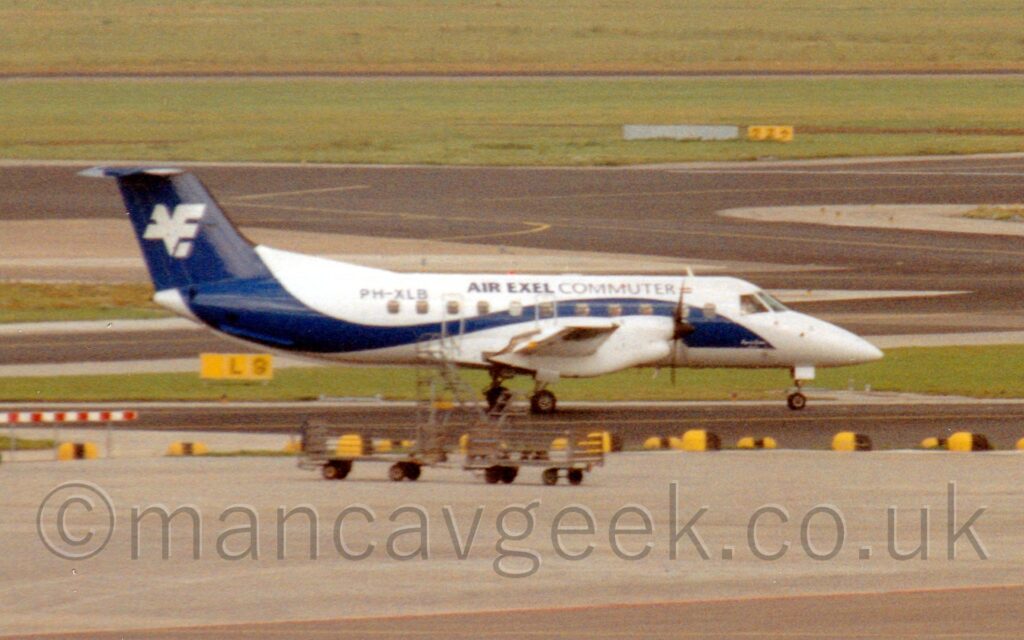 Somewhat grainy, slightly soft photo of a twin propellor-engined airliner taxiing from left to right along a black taxiway. The plane is mostly white, with a dark blue stripe swooping its way from the lower forward fuselage, over the wings, then up to fill the tail. There are black "Air Exel Commuter" titles on the upper forward fuselage, with the black registration "PH-XLB" on the upper rear. A white logo that seems to combine the letters "X" and "E" is on the tail. Grey concrete apron fills the foreground, with strips of grass lining several black taxiways that wander through the background, with more grass in the distance.