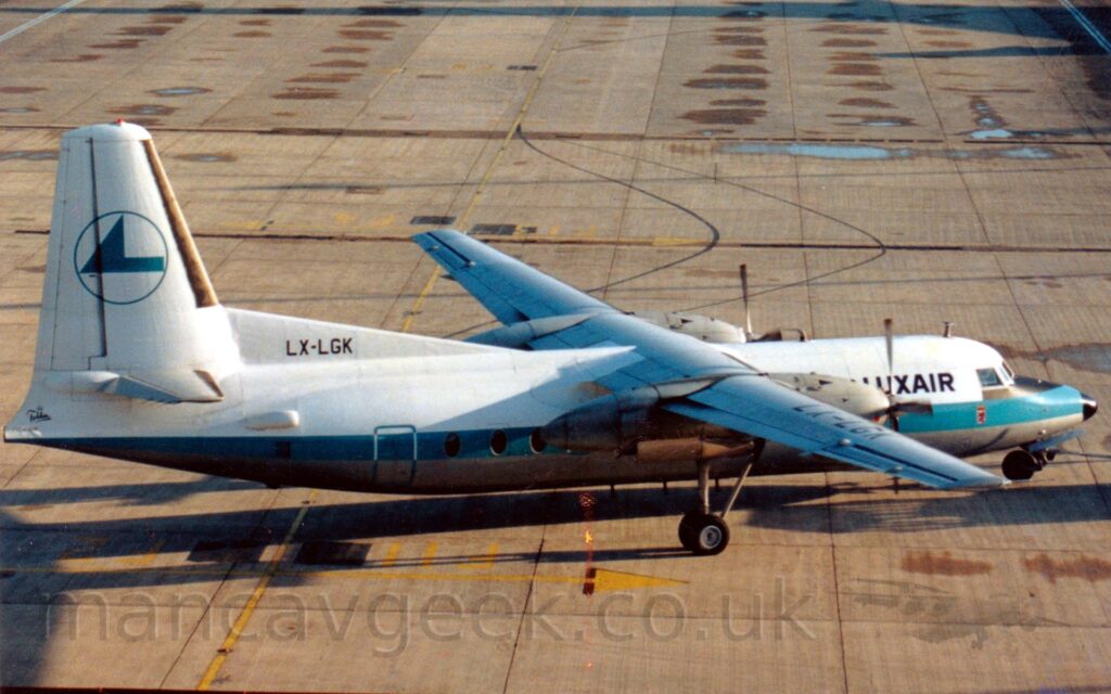 High side view of a high-winged, twin propellor-engined airliner facing to the right but being pushed back to the left by an out-of-frame tug attached to the nose-wheel by a long blue pole. The plane is mostly white, with a grey belly, and a thick, light blue stripe running along the body. There are black "LuxAir" titles on the upper forward fuselage, between the cockpit windows and the wing. The tail is white, with the blue outline of a circle containing a rather angular, blue, stylised flying bird, and the black registration "LX-LGK" on a fillet at the front. Grey concrete apron with occasional damp patches fill the rest of the frame.