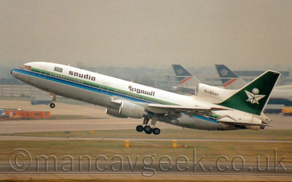 Side view of a 3 engined jet airliner flying from right to left at a very low altitude, just metres above a black runway, with undercarriage lowered, flaps extended from the front and rear of the wings, and the nose raised sharply, suggesting it has just taken off. The plane is largely white, with a green and blue stripe running along the body, covering the passenger cabin windows, and a grey belly. There are black "Saudia" titles on the upper forward fuselage, next to a green flag covered in white Arabic writing. The Saudia titles are repeated on the upper mid-fuselage, over the wing root, this time in an Arabic script. There is a small, black number "5" on the lower part of the nosecone. There is black text "TriStar" on the centre engine inlet, on the upper rear fuselage, in front of the tail. The tail is green, outlined in white, with green crossed swords over an inverted white triangle, with a white palm tree at the top. A black runway stretches across the bottom of the frame, lined with grass on either side. A couple of very large jet airliners with dark blue and grey tails can be seen in the background, on the right, under grim grey sky.