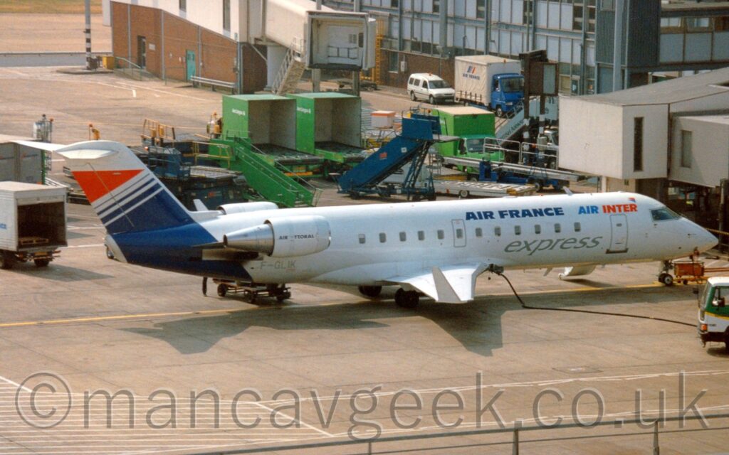 Side view of a twin-engined jet airliner parked facing to the right, nose-in to a terminal building. The plane is mostly white, with dark blue "Air France" and light blue and red "Air Inter" titles on the upper forward fuselage, with grey "Express" titles on the belly. The engines, mounted on the sides of the rear fuselage, have black "Air Littoral" titles on the sides. The rear fuselage and tail is mostly dark blue, with blue stripes and a red triangle at the top, all outlined with white. Grey concrete apron fills the foreground, with green vans, white vans, and blue baggage trucks and airstairs scattered around the background, with a terminal building behind all those at the top of the frame.