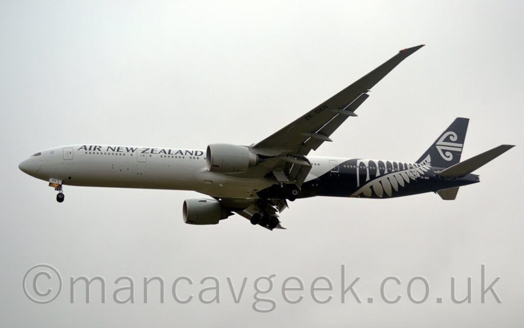 Side view from below of a twin engined jet airliner flying from left to right at a low altitude, with undercarriage lowered and flaps extended from the front and rear of the wings, suggesting the it is about to land. The plane is mostly white, with black "Air New Zealand" titles on the upper forward fuselage, while the rear fuselage is black, with white fern leaves, and stylised white waves on the black tail, with the white registration "ZK-OKN" on the lower rear fuselage. Flat white sky fills the rest of the frame.