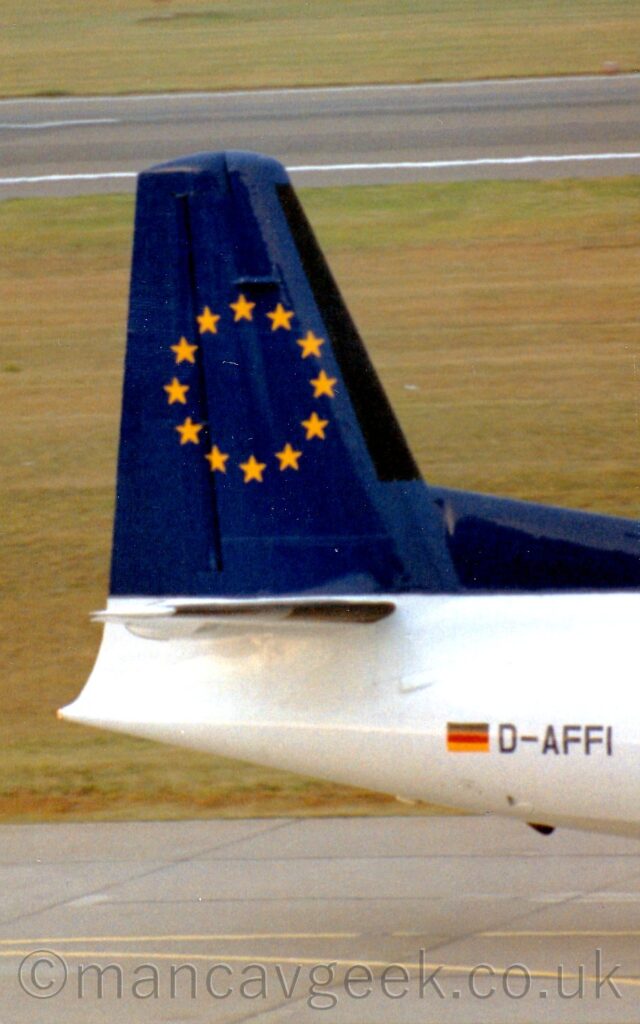 Close-up side view of the tail of an airliner taxiing from left to right. The plane's body is almost entirely white, with the black registration" D-AFFI" next to a small German flag on the rear fuselage. The tail is dark blue, with a circle of 12 golden stars in the middle. Grey concrete taxiway is visible at the bottom of the frame, while large patches of grass fill the background, divided by a black runway in the distance.