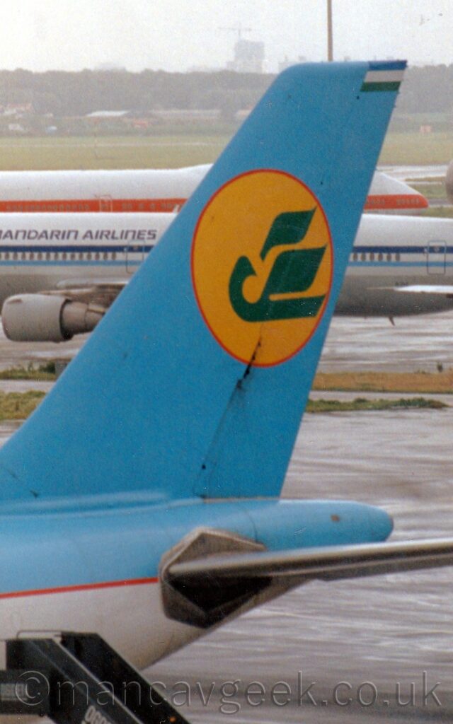 Close-up of the rear fuselage and tail of a jet airliner parked facing to the left. The plane is mostly white, with a blue top section, separated by a thin red stripe. The tail is the same blue, with a yellow circle outlined in red, with a stylised green flying big bird in the centre. Grey concrete taxiways fill the background, with a couple of thin strips of grass, and a couple of white jet airliners, one with a blue stripe, the other with a red stripe. More grass beyond them leads up to trees and buildings in the distance, slowly vanishing in to the hazy grey sky.