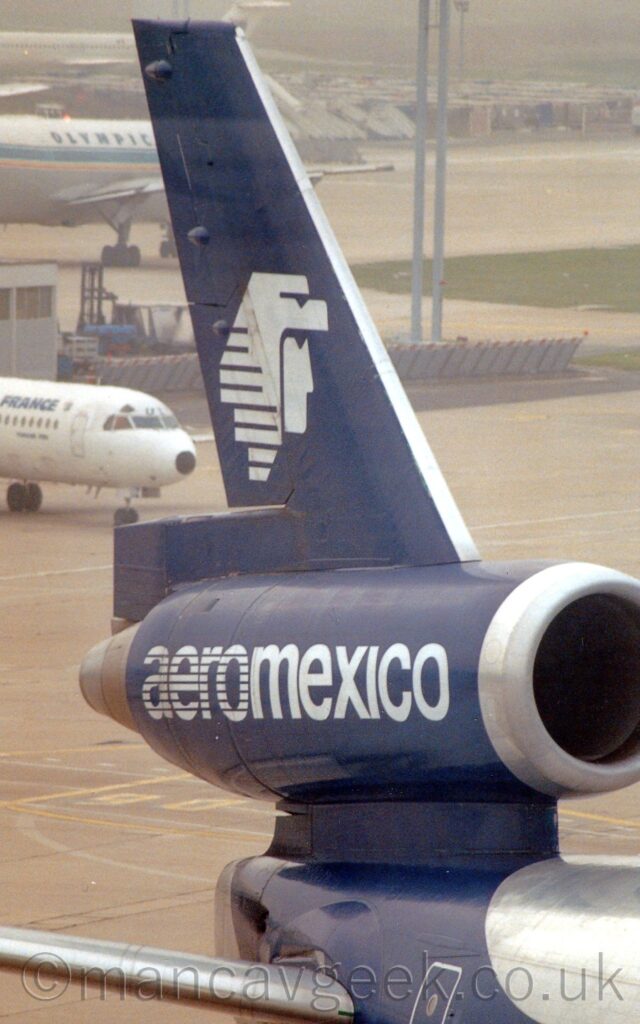 Close-up of the rear fuselage, centre engine, and tail of a 3 engined jet airliner parked facing to the right. There is a dark blue diagonal band wrapped around the rear fuselage and tail, with white "AeroMexico" titles on the sides of the engine pod that runs straight through the tail, and the stylised white head of a person facing to the right who is wearing an animals head as a head covering. Grey concrete apron extends into the distance in the background, with the forward fuselage of a small, white jet airliner poking into frame on the left, and the rear fuselage of a larger jet airliner taxiing past behind a grey building further back on the left.