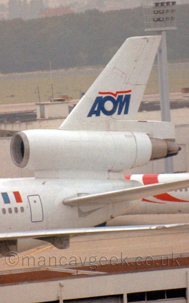 Close-up of the rear fuselage and tail of a 3 engined jet airliner taxiing from right to left. The plane is almost entirely white, with a large French flag over the rear passenger cabin windows. The tail has the dark blue letters "AOM", with a stylised red flying bird over the top, made up of a single line. The roof of a small building stretches across the frame in the foreground, with another building stretching across the background, this one with a smaller white and red airliner parked in front. A large expanse of grass beyond that leads up to trees in the distance.