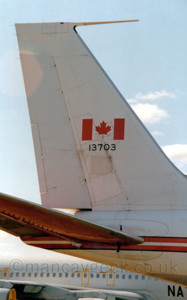 Close-up of the white rear fuselage and tail of a military transport plane. The plane has a large Canadian flag (red field either side of a larger white field containing a red maple leaf) in the centre of the tail, over the black serial "13703". There is a red-white-red stripe, outlined in black, running along the body. In the background, a grey, 4 jet-engined military plane with the black letters "NAT" on the lower forward fuselage can be seen at the bottom of the frame. The rest of the background is pale blue sky with fluffs of white cloud.