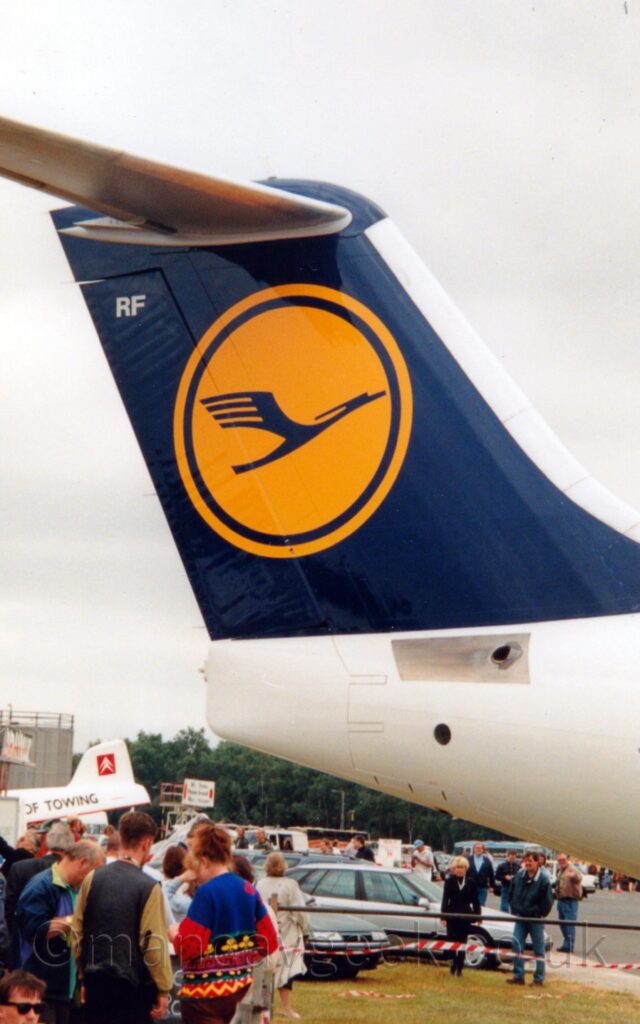 Vertically-oriented shot of the rear fuselage and tail of a jet airliner parked facing to the right. The fuselage is white, while the tail is deep blue, with a yellow circle containing a stylised image of a dark blue flying bird. A group of people can be seen in the background under the planes tail, with cars, buildings, and trees in the distance, under flat grey sky.