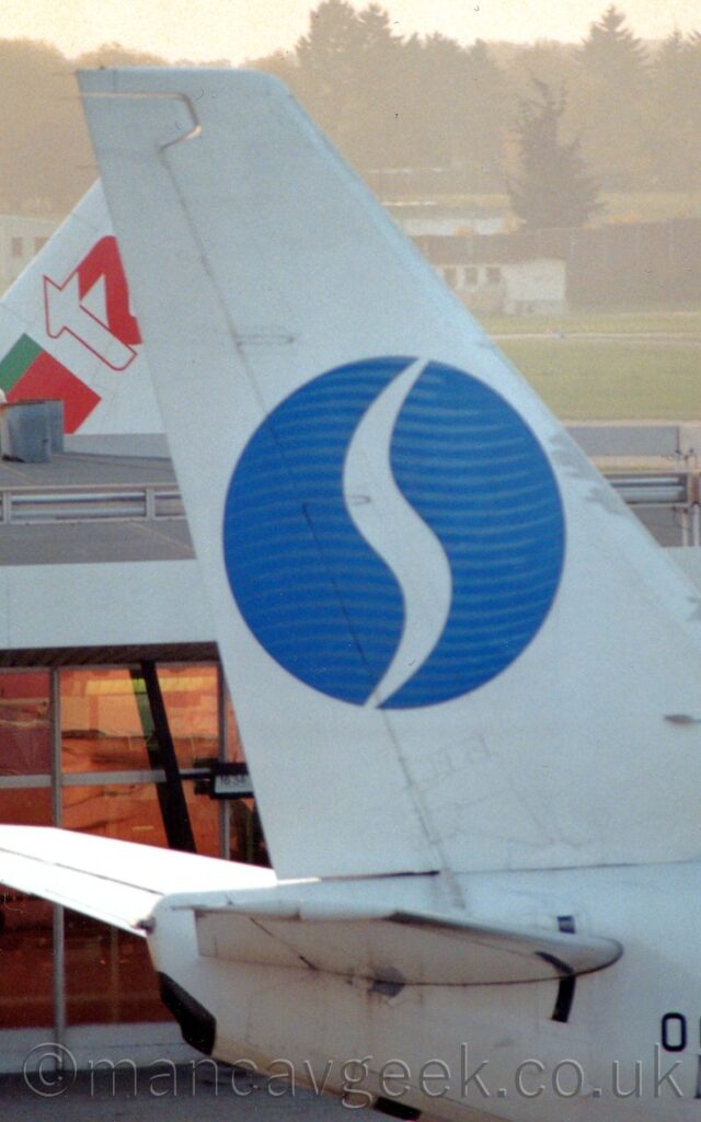 Close-up of the tail of a jet airliner parked facing to the right. The plane is mostly white, with a blue circle in the middle, with pale blue bands giving the impression of a globe, overlaid with a tall, skinny, stylised letter "s". A heavily glazed grey terminal building fills the background, with the white, red, and green tail of another airliner beyond. Grass in the background leads up to white buildings and trees in the distance, slowly being swallowed up by bright, hazy sky.