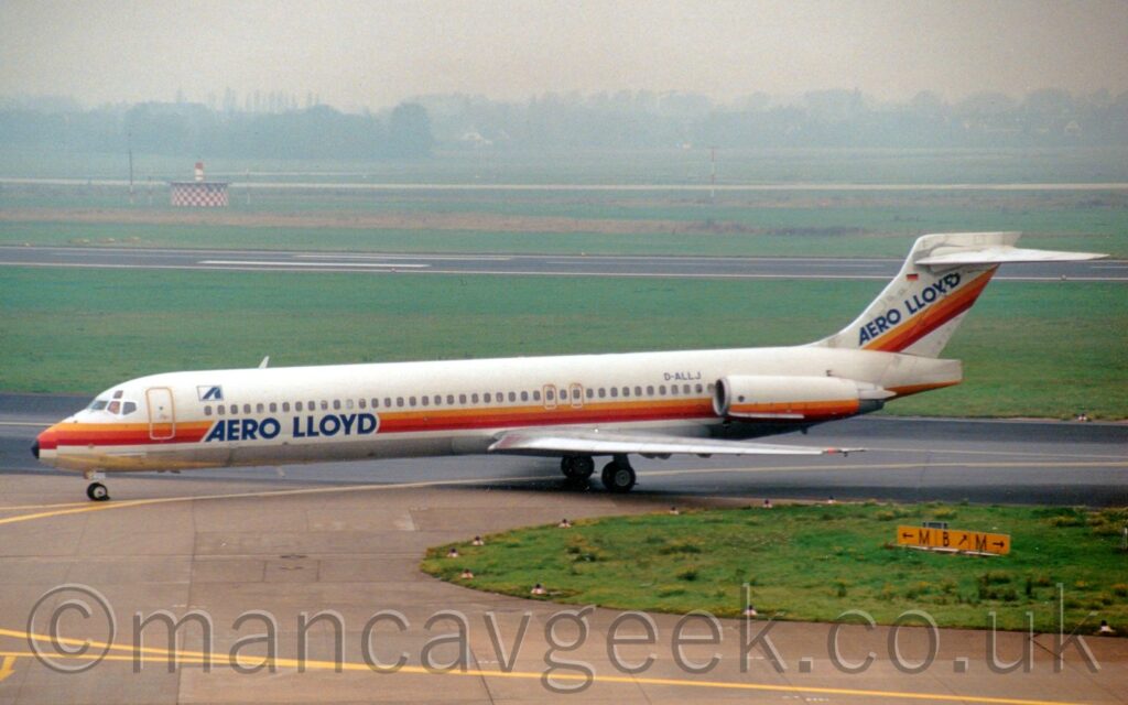 Side view of a twin engined jet airliner, with the engines mounted on the sides of the rear fuselage, taxiing from right to left following a yellow line marked on the taxiway. The plane is white, with an orange and yellow stripe running along the body under the passenger cabin windows, overlaid with dark blue text "Aero Lloyd" on the lower forward fuselage. The stripe also runs along the bottom of the engine pods, as well as diagonally across the tail, under blue "Aero Lloyd" titles. The black registration "D-ALLJ" is on the upper rear fuselage, just in front of the engines. Grey taxiway fills the bottom of the frame, with a strip of grass coming from the right separating that from a darker grey taxiway that the plane is taxiing off. More grass in the background separates the taxiways from a grey runway, with more grass leading up to trees in the distances, slowly vanishing into hazy grey clouds.