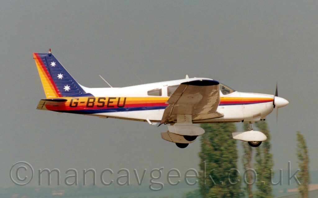 Side view of a single-engined light aircraft flying from left to right at a low altitude, with flaps fully extended from the rear of the wing, suggesting it is about to land. The plane is largely white, with a yellow, orange, red, and dark blue stripe running along the body, getting wider as it moves rearwards, overlaid with the black registration "G-BSEU" on the rear fuselage. The stripes are reversed and arranged vertically on the tail - dark blue at the front, thin thinned red, orange, and yellow stripes, with 3 white stars in the blue part. White spats cover the fixed undercarriage under the wings and the nose. Grey hangars and tall trees fill the lower part of the background, with grey sky filling the rest.