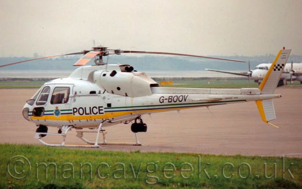 Side view of a white helicopter parked on grey concrete apron, facing to the left. A yellow stripe runs along the bottom of the body, as well as along the leading edge of the tail, above and below the end of the tail boom. Green and blue stripes run along the body a bit further up, overlaid by black "Police" text just aft of the cockpit doors, with the badge of the Merseyside Police on the door.. The black registration "G-BOOV" is on the top of the tail boom. Green grass fills the foreground, with grey apron filling at lo of the background, with more grass leading up to a wide river, with fields and trees on the far side. Grey sky fills the rest of the frame.