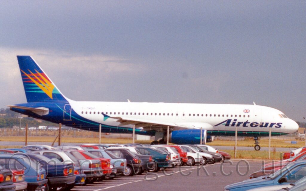 Side view of a twin engined jet airliner taxiing from left to right along a grey taxiway. The plane is mostly white, with a blue belly, sweeping up through the rear fuselage to fill the tail, accompanied by a thin green stripe. There are large, dark blue "AirTours" titles on the lower forward fuselage, with a small UK flag above the cabin windows, above the letter "t". The dark blue tail has 3 sets of 3 long, thin triangles, red and the top and green at the bottom, overlaid with yellow diagonal triangles. A ground level car park fills the foreground, with plenty of colourful cars, with a chainlink fence between the cars and the plane. Grey sky fills the background.