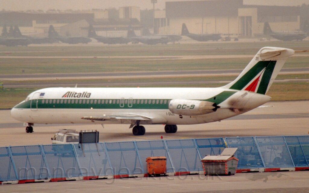 Side view of a twin engined jet airliner taxiing from right to left and slightly away from the camera. The plane is largely white, with a thick green stripe running along the body, covering the passenger cabin windows, with black "Alitalia" titles on the upper forward fuselage, with a red triangle in the middle of the initial letter "A". A similar letter "A", this time in green, fills the white tail. The engine pods, mounted on the sides of the rear fuselage, has the text "DC-9" in green. The black registration "I-DIZE" is on the lower rear fuselage, just in front of the engines, with the last 2 letters "ZE" repeated at the top of the tail and on the open nose wheel doors. A blue metal mesh blast barrier stretches across the foreground, with red and white striped blocks marking it's position for planes, and a couple of large dumpster bins in front. Alternating strips of grass and taxiways and runways lead off into the distance, with a collection of high-winged, green or grey military transport aircraft on the far side, in front of cream-coloured buildings, under grim grey sky.