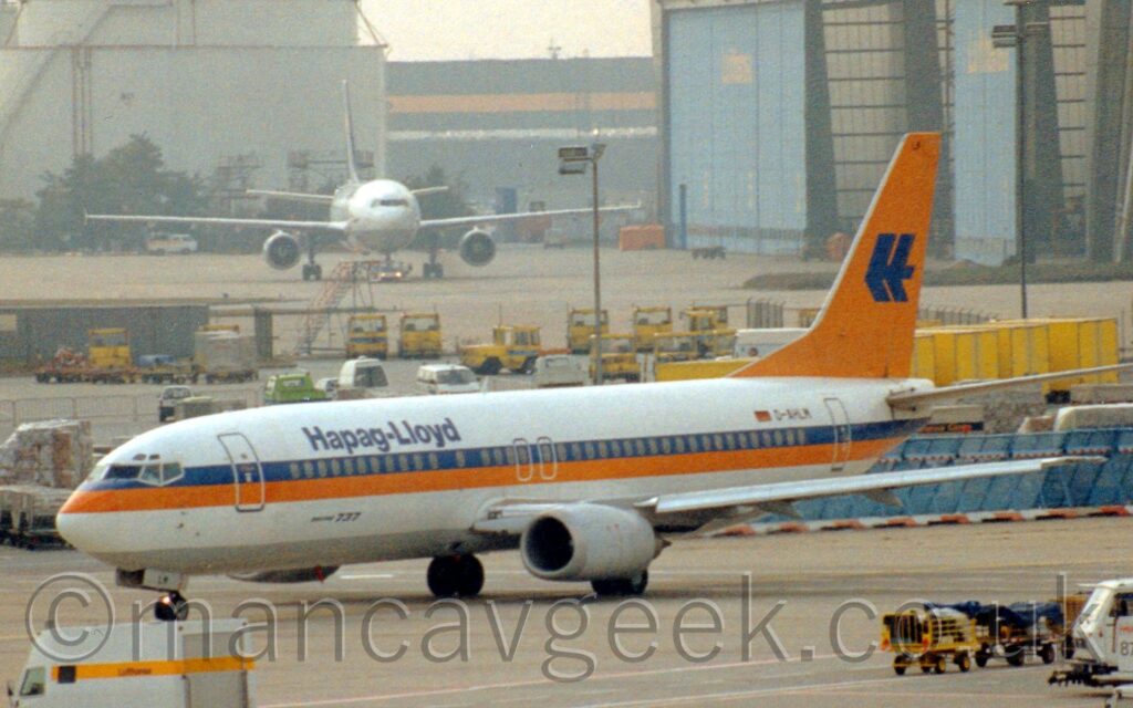 Side view of a twin engined jet airliner taxiing from right to left. The plane is mostly white, with a thick blue and orange cheatline running along the body from the nose, the blue part covering the passenger cabin windows. There are blue "Hapag-Lloyd" titles on the upper forward fuselage, and the registration "D-AHLM" next to a small German flag on the upper rear. The letters "LM" are on the open nose-wheel doors, and at the top of the orange tail. The tail also has a large stylised version of the letters "HL" in the middle. Grey concrete apron fills the foreground, with white trucks and yellow baggage carts driving around at the edges, with more yellow baggage trucks parked in the background. In the distance, a large grey hangar with pale blue doors is on the right of the frame, with a white, twin engined airliner parked outside, more or less facing the camera, with large pale blue fuel tank on the left.