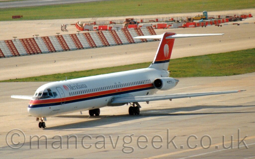 Side view of a twin engined jet airliner with the engines mounted on the sides of the rear fuselage, taxiing from right to left along a grey taxiway. The plane is largely white, with a red and black stripe with a gold pinstripe along the middle running along the body under the passenger cabin windows. There are black "Meridiana" titles on the upper forward fuselage, just aft of a red circular logo. The tail is white at the base, with most of the rest being red, with a larger version of that circular logo, this time in white, with a black stripe and yellow pinstripe at the bottom. The white engine pods have black text "DC-9-51" on the side. Grey concrete apron fills the foreground, light coming from the top left corner casting a strong shadow of the plane on the ground. A strip of green grass on the left of the background separates this plane from a couple of taxiways in the background, the taxiways themselves being separated by ay red and white striped metal fence coming from the left, and a small building site behind red alternating red and white barricades, with more grass at the top of the frame.