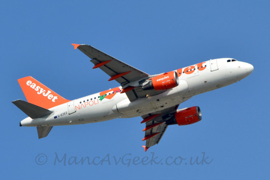 Low side view of a white, twin engined jet airliner flying from left to right at a very low altitude. It's undercarriage is already tucked away, it's flaps are extended behind the wings, and it's nose is raised sharply, suggesting that it has just taken off. The word "Jet" is visible in large orange letters on the forward fuselage, with a large, orange love heart and the word "Napoli" on the rear fuselage. The engine pods slung under the wings are bright orange, with the word "EasyJet" in white, a design replicated on the tail. The registration "G-EZEZ" is on the lower rear fuselage in black, repeated under the left wing. Under the wings, the covers on the flap tracks are bright orange, as are the wing-tips. Pale blue sky fills the rest of the frame.