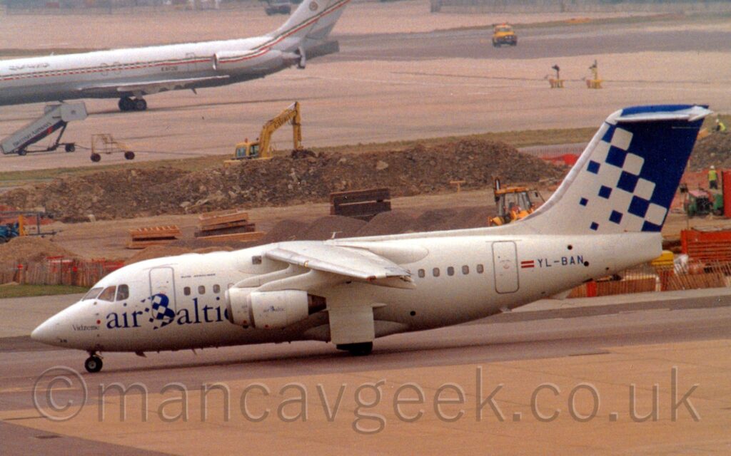 Side view of a high-winged, 4 engined jet airliner taxiing from right to left along a grey taxiway. The plane is mostly white, with blue "Air Baltic" titles on the lower forward fuselage, and the dark blue registration "YL-BAN" next to a small Lithuanian flag on the side of the rear fuselage. The tail starts off white a the front, with blue squares slowly taking over and merging to fill the rear of the tail. A building site is in the background, with large mounds of dirt and rubble, and various yellow diggers scattered around. In the distance, a twin engined jet airliner is on the left of the frame, it's nose out of shot, with more concrete apron filling the rest.