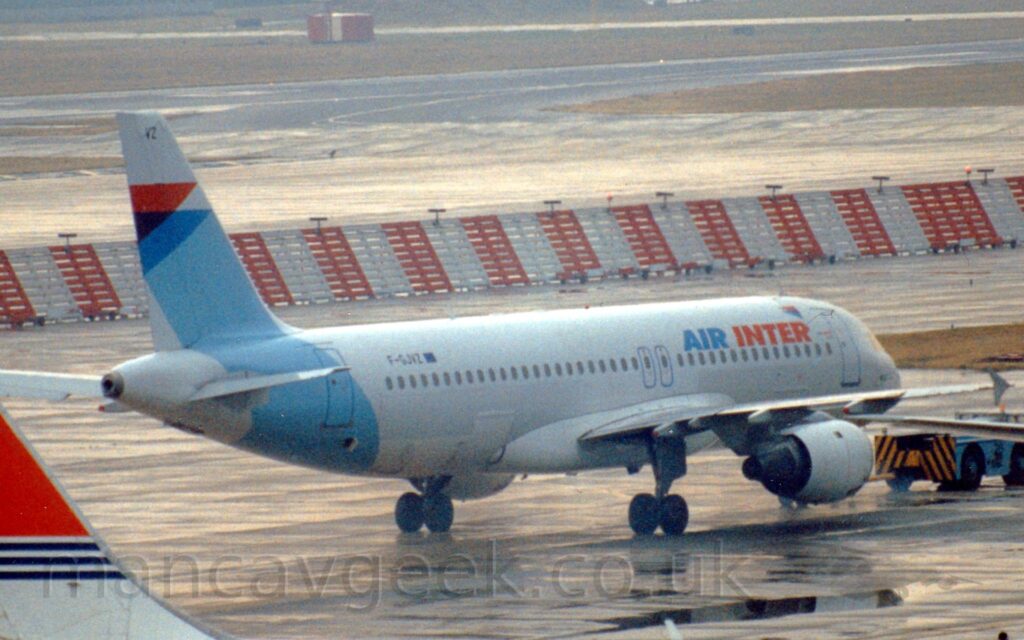 Side view of a twin engined jet airliner facing to the right but being pushed back to the left by a low-slung blue tuk with a black and yellow chevron pattern on the back. The plane is mostly white, with a diagonal pale blue band around the rear fuselage and tail, with red and darker blue triangles at the top, and red and dark blue "Air Inter" titles on the upper forward fuselage. Damp grey apron fills the foreground, with part of the red and white tail of another jet airliner in the bottom left corner, and a red and white striped metal blast fence running across the frame in the background, with a grey taxiway and black runway lined with grass at the top of the frame.