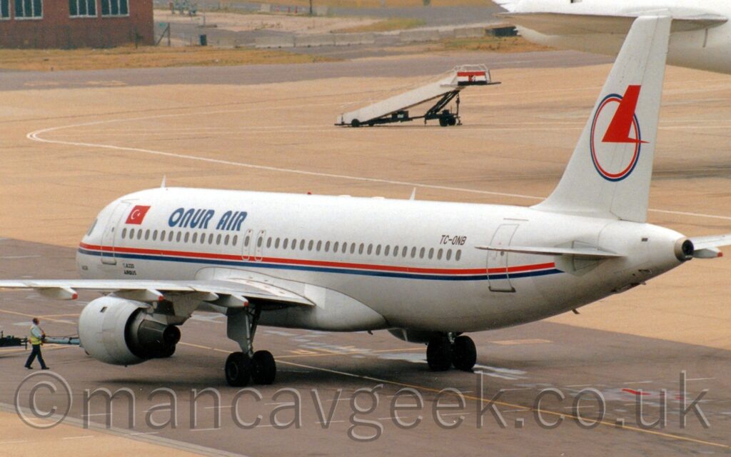 Side view of a twin engined jet airliner , facing to the left but being pushed back to the right by an out of frame tug attached to the nose-wheel by a long white pole. The plane is largely white, with a thin red and blue stripe running along the body under the passenger cabin windows, and dark blue "Onur Air" titles on the upper forward fuselage. There is a red and white Turkish flag just aft of the forward cabin door., and the black registration "TC-ONB" on the upper rear fuselage. The tail is white, with the blue and red outline of a circle, overlaid with a stylised, red, flying bird in the top right corner. There is a man wearing a yellow hi-viz vest walking alongside the plane as it is pushed back, with grey concrete apron filling the rest of the foreground. More apron fills the background, with the all-white rear fuselage and tail of another jet airliner in the top right corner, and a low brown brick building in the top left.