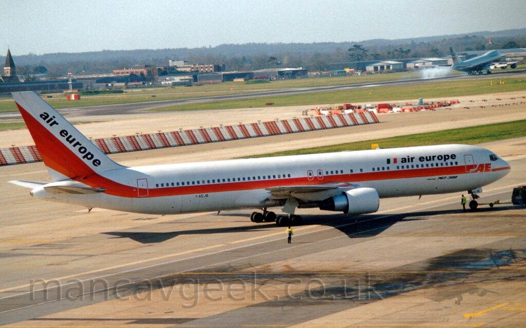 Side view of a twin engined jet airliner facing to the right but being pushed back to the left by an almost-out-of-frame blue tug attached to the nosewheel by a long blue pole. The plane is mostly white, with a red and orange stripe running along the body below the passenger cabin windows, before sweeping up into the tail. There are black "Air Europe" titles next to an Italian flag on the upper forward fuselage, as well as diagonally on the white tail. Yellowing grey concrete apron fills the foreground, with strips of grass, taxiways, and a black runway in the background, with a very large, 4 engined jet airliner just touching down on the runway in the top right corner. Cargo sheds, office buildings, and a church are nestled in trees in the distance, under hazy blue-grey sky.