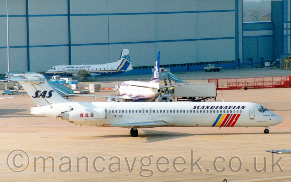 Side view of a white, twin engined jet airliner with the engines mounted on the side of the rear fuselage, taxiing from left to right. There are a number of coloured near-vertical bars - blue, yellow, red, and black - on the lower forward fuselage, starting under the passenger cabin windows and looping under the belly, as well as squares on the engine pods - red with a white cross, red with a black cross inside a white cross, and blue with a yellow cross, each representing the countries Sweden, Norway, and Denmark. There are black "Scandinavian" titles on the upper forward fuselage and a black "SAS" logo on the tail. Grey concrete fuselage fills the foreground, with another white, twin engined jet airliner with a blue belly and tail in the immediate background, facing more or less away from the camera. Beyond that, a white, twin propellor-engined airliner with a blue belly and a yellow stripe running along the body is parked facing to the left in front of a large grey hangar, the hangars doors dwarfing this latter plane.