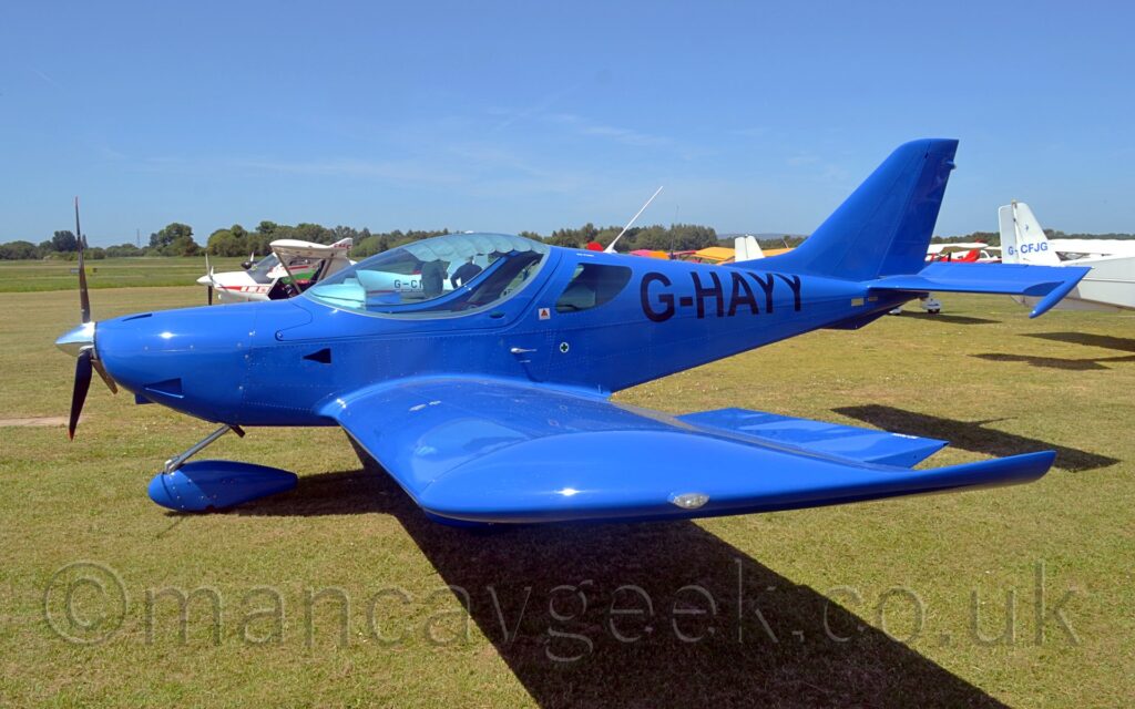 Side view of a single engined light aircraft parked on grass facing to the left. The plane is a vivid blue, with black registration "G-HAYY" on the rear fuselage. A large bubble canopy covers the 2 seat cockpit. The plane is casting a strong shadow almost straight down on the ground, suggesting it is sunny and the sun is almost overhead. A collection of light aircraft are parked behind this plane, with trees lining the airfield perimeter in the distance, with bright blue sky filling the rest of the frame.